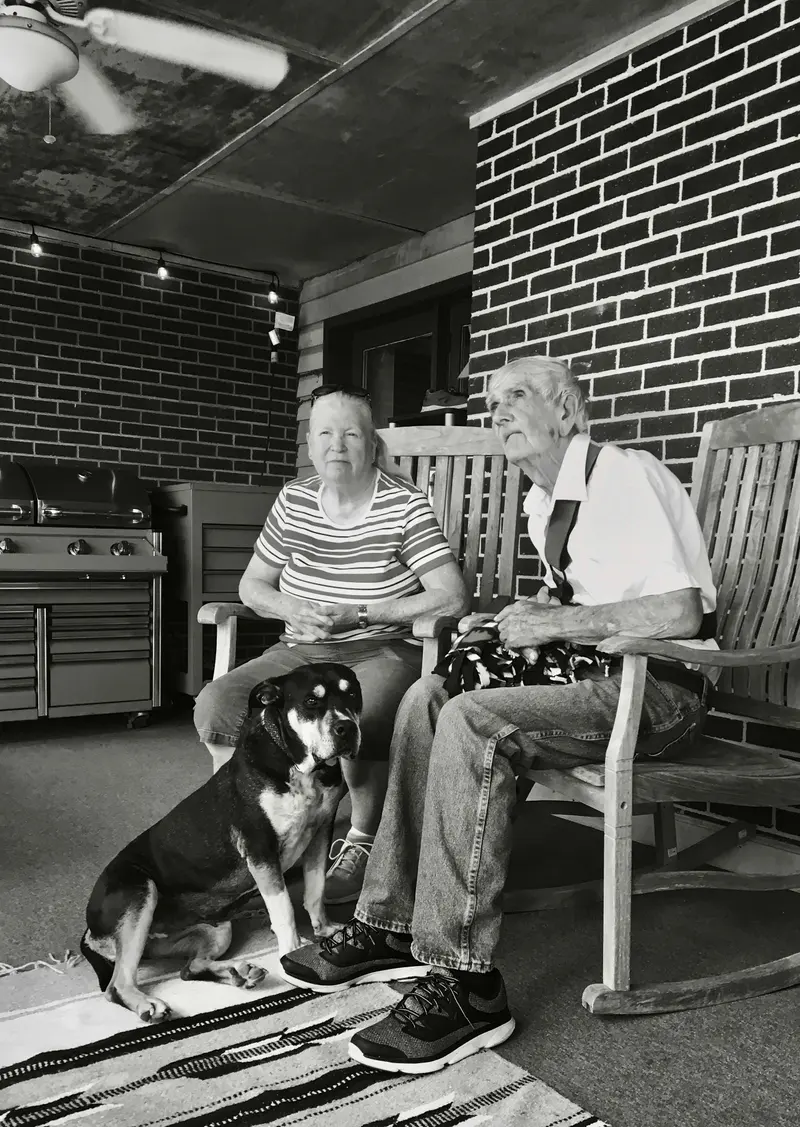 An elderly couple in rocking chairs on a porch with their grey-muzzled dog sitting at their feet, black and white